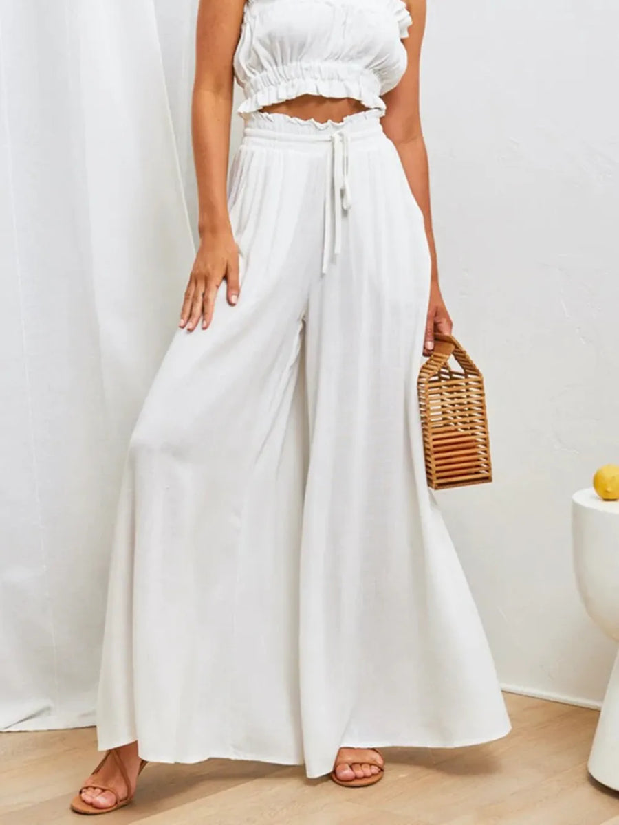 Person wearing a white outfit with wide-leg pants and a woven bag, standing against a plain background.