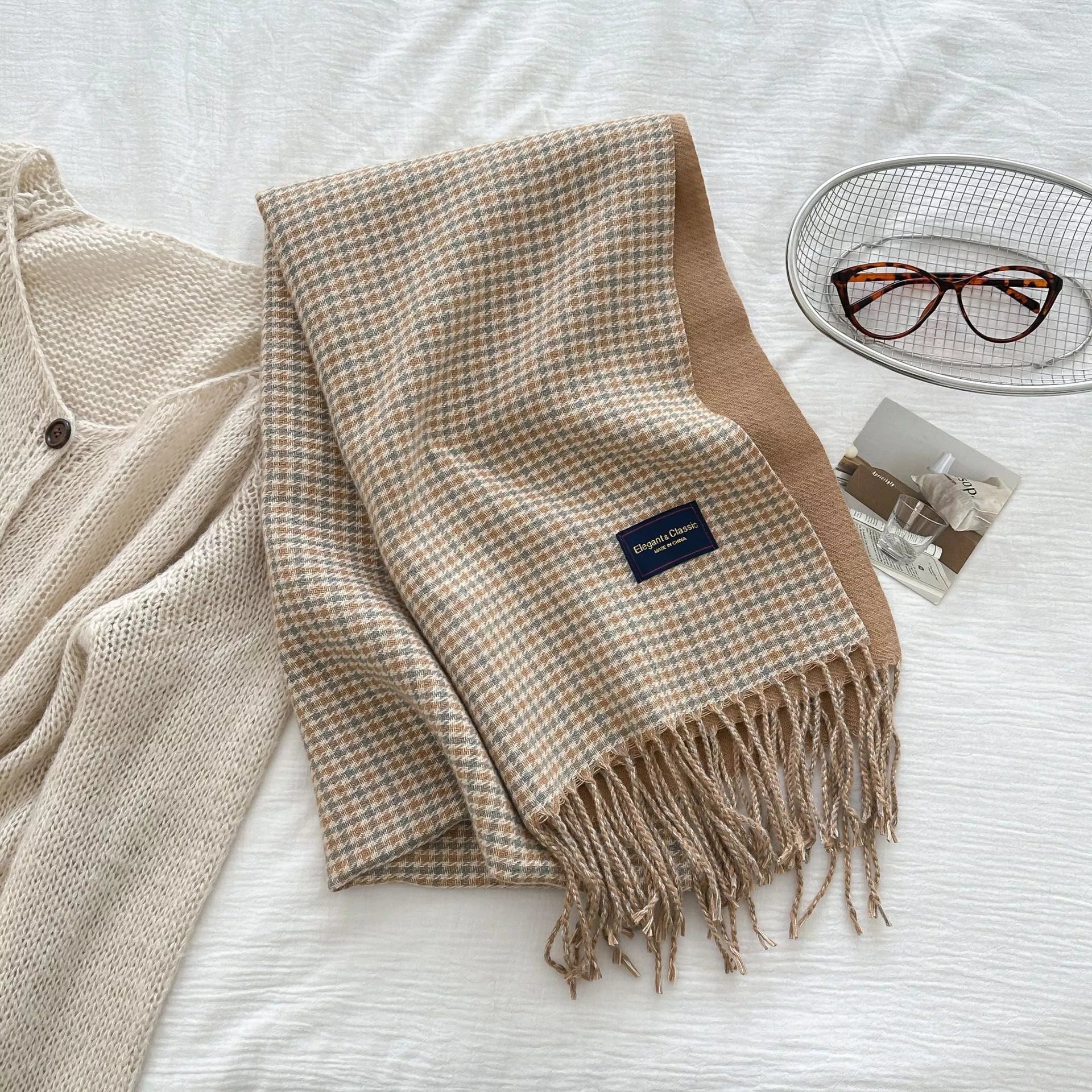 Beige plaid scarf with a brand label on a white surface, next to a beige cardigan and glasses.