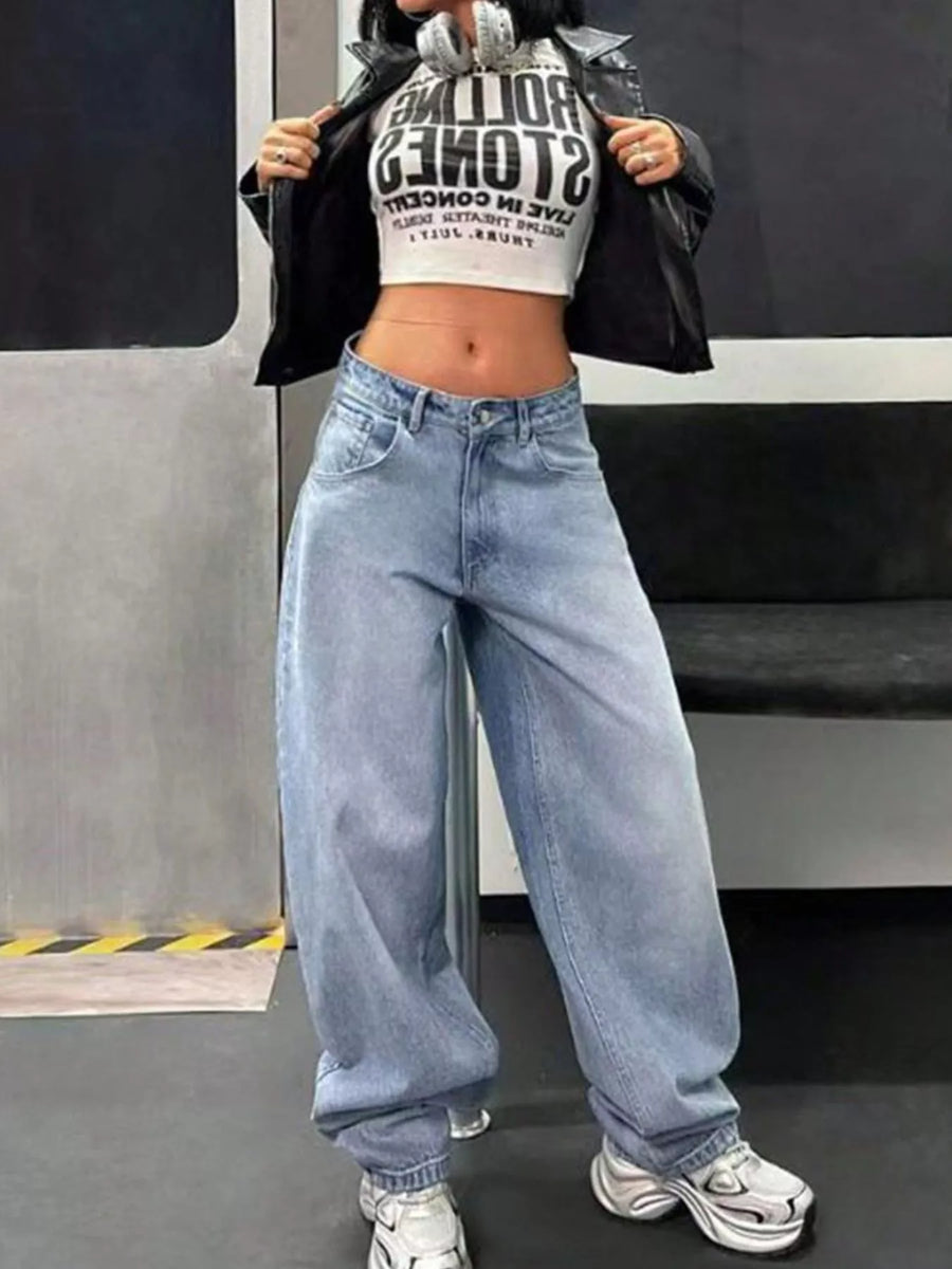 Person wearing a black jacket, white crop top with text, and blue jeans standing in front of a train.