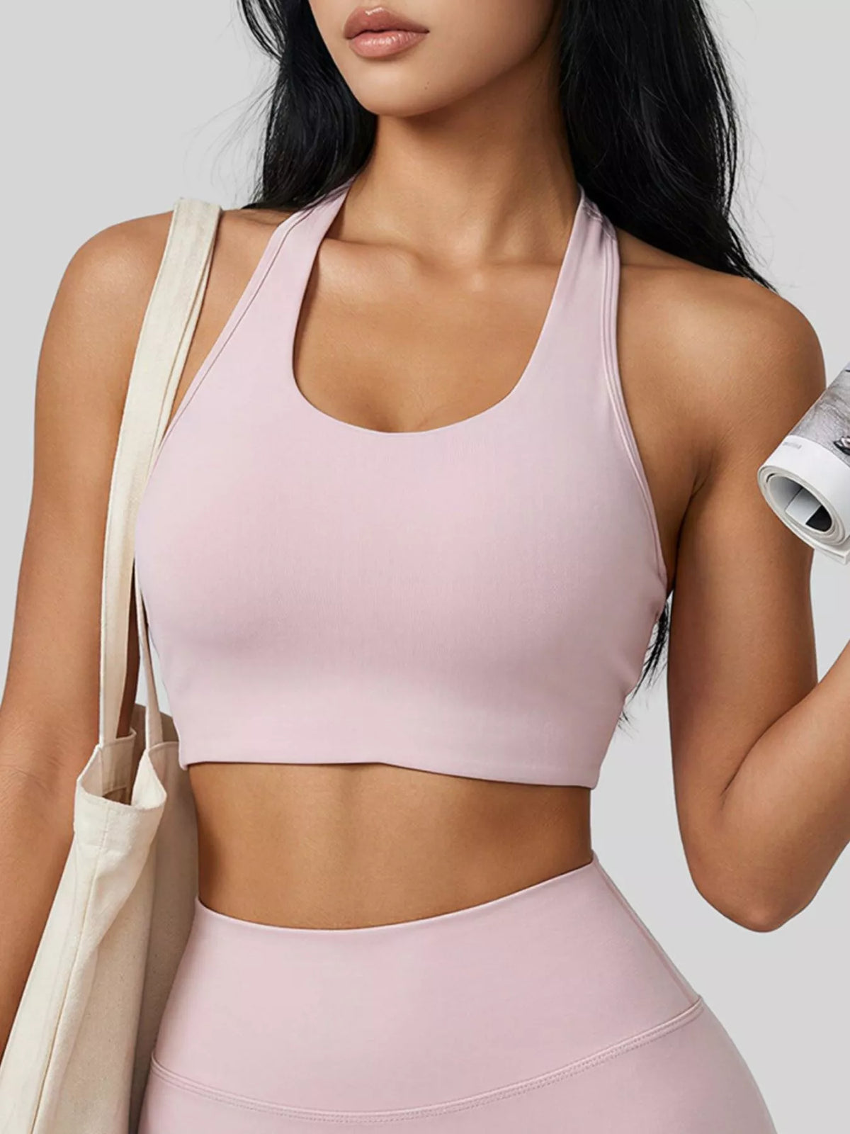 A woman wearing a light pink athletic sports bra and matching high-waisted leggings, carrying a cream canvas tote bag against a light gray studio background.
