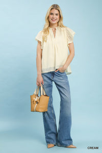 Woman wearing a cream blouse and blue jeans, holding a tan handbag on a light blue background.