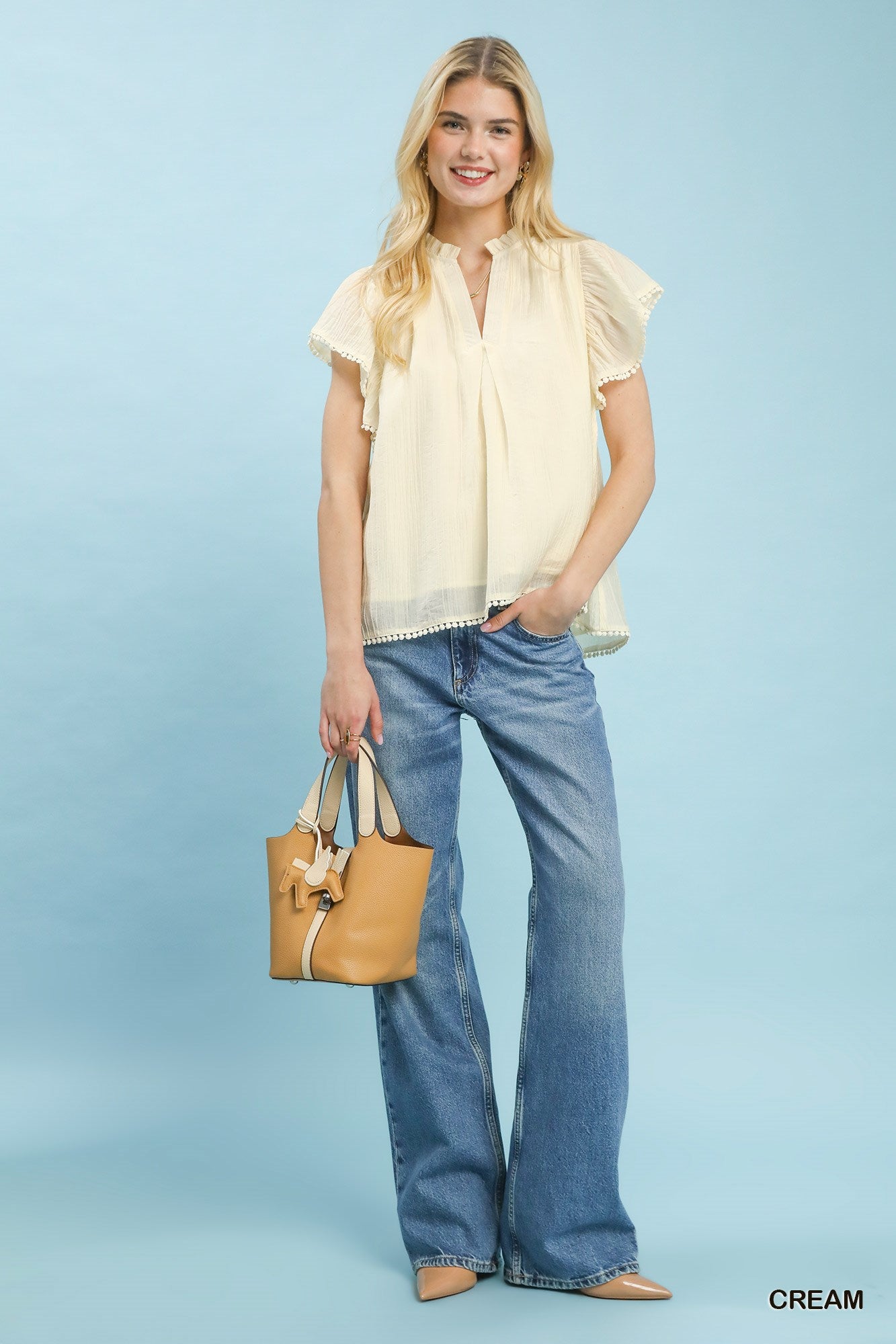 Woman wearing a cream blouse and blue jeans, holding a tan handbag on a light blue background.