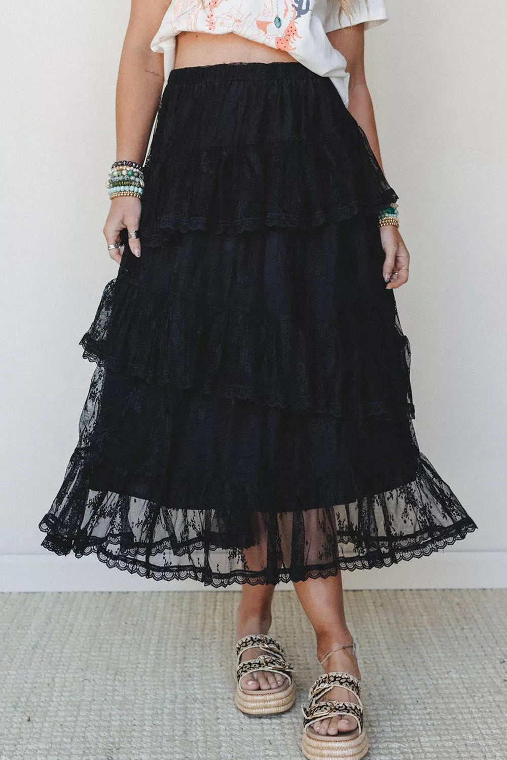 A black tiered tulle maxi skirt worn by a model, paired with a white floral crop top and brown espadrille sandals, standing against a plain white wall.