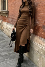 A woman wearing a long-sleeve brown midi dress with an asymmetrical draped hem and side ruching, paired with black knee-high boots and holding a black handbag, standing against a red brick wall.