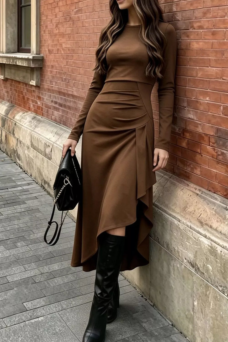 A woman wearing a long-sleeve brown midi dress with an asymmetrical draped hem and side ruching, paired with black knee-high boots and holding a black handbag, standing against a red brick wall.