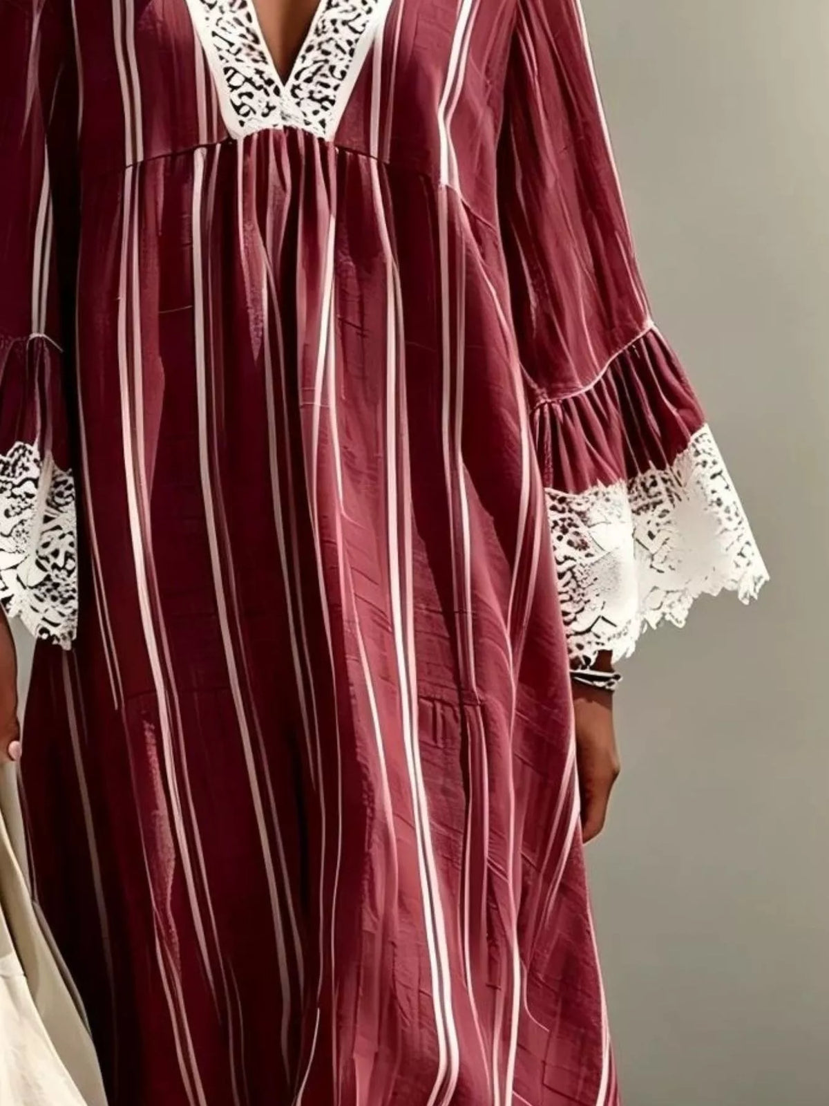 A close-up of a long-sleeved dress or tunic in deep burgundy with vertical white stripes and white lace trim at the V-neck and cuffs, shown on a model against a plain gray background.