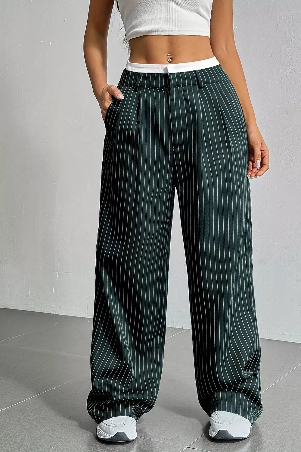 A person wearing dark green pinstriped wide-leg pants with a white waistband, paired with a white crop top and white sneakers, standing against a light gray wall.