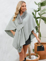 Woman wearing a gray poncho with fur trim standing next to potted plants.
