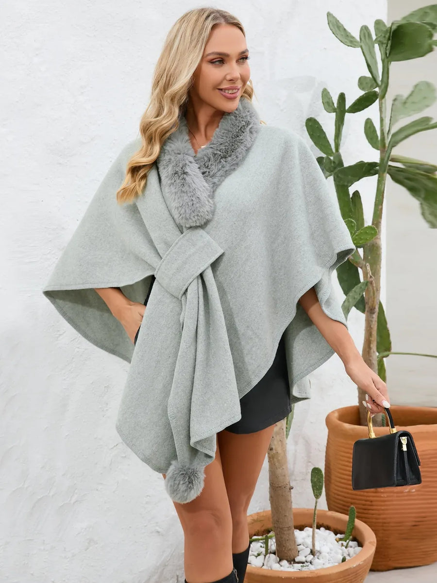 Woman wearing a gray poncho with fur trim standing next to potted plants.