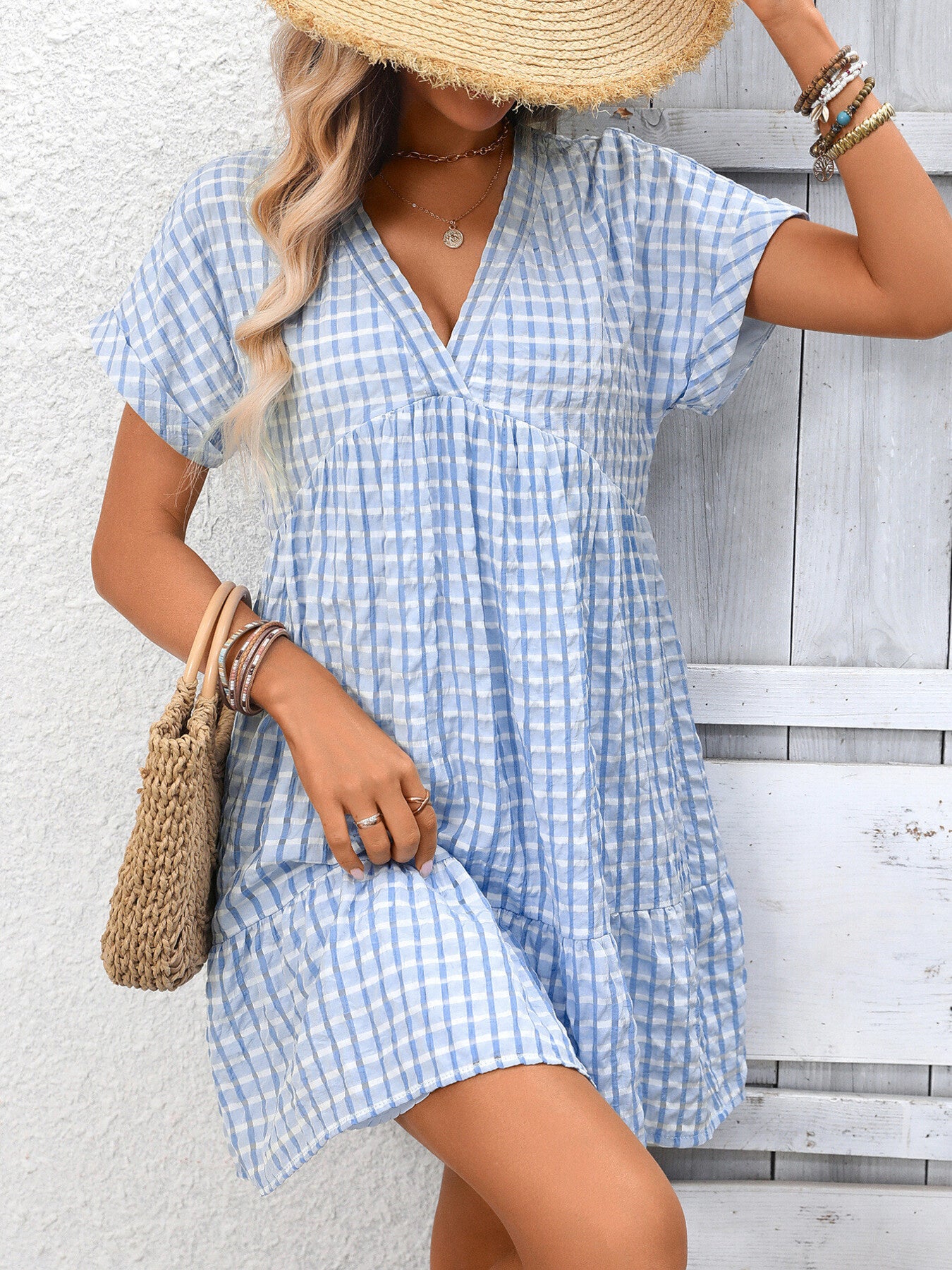 Woman wearing a blue and white checkered dress with a straw hat and holding a woven bag against a light wooden background.