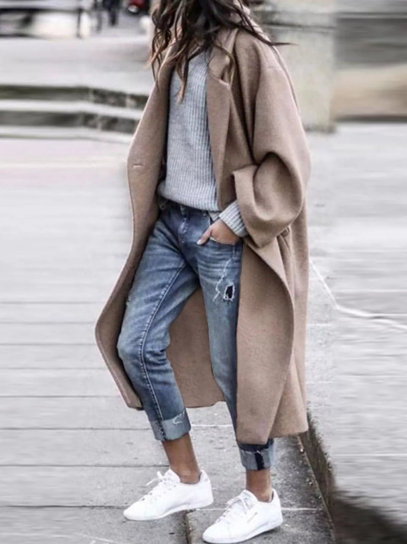 Person wearing a beige coat, light blue jeans, and white sneakers on a blurred street background