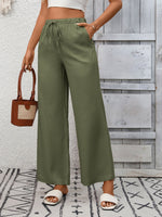 Person wearing green wide-leg pants with a white top and sandals, holding a brown handbag on a patterned rug.