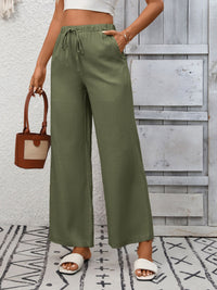 Person wearing green wide-leg pants with a white top and sandals, holding a brown handbag on a patterned rug.