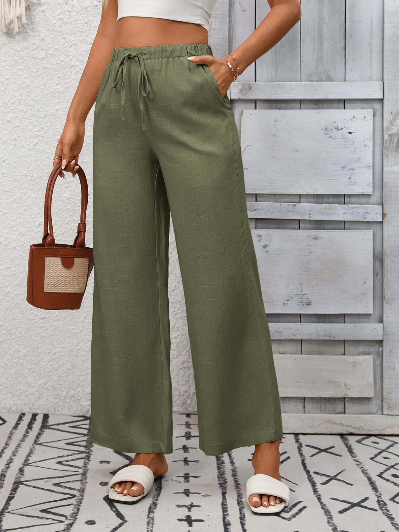 Person wearing green wide-leg pants with a white top and sandals, holding a brown handbag on a patterned rug.