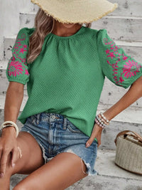 Woman wearing a green top with pink floral patterns and denim shorts, sitting on a wooden floor.