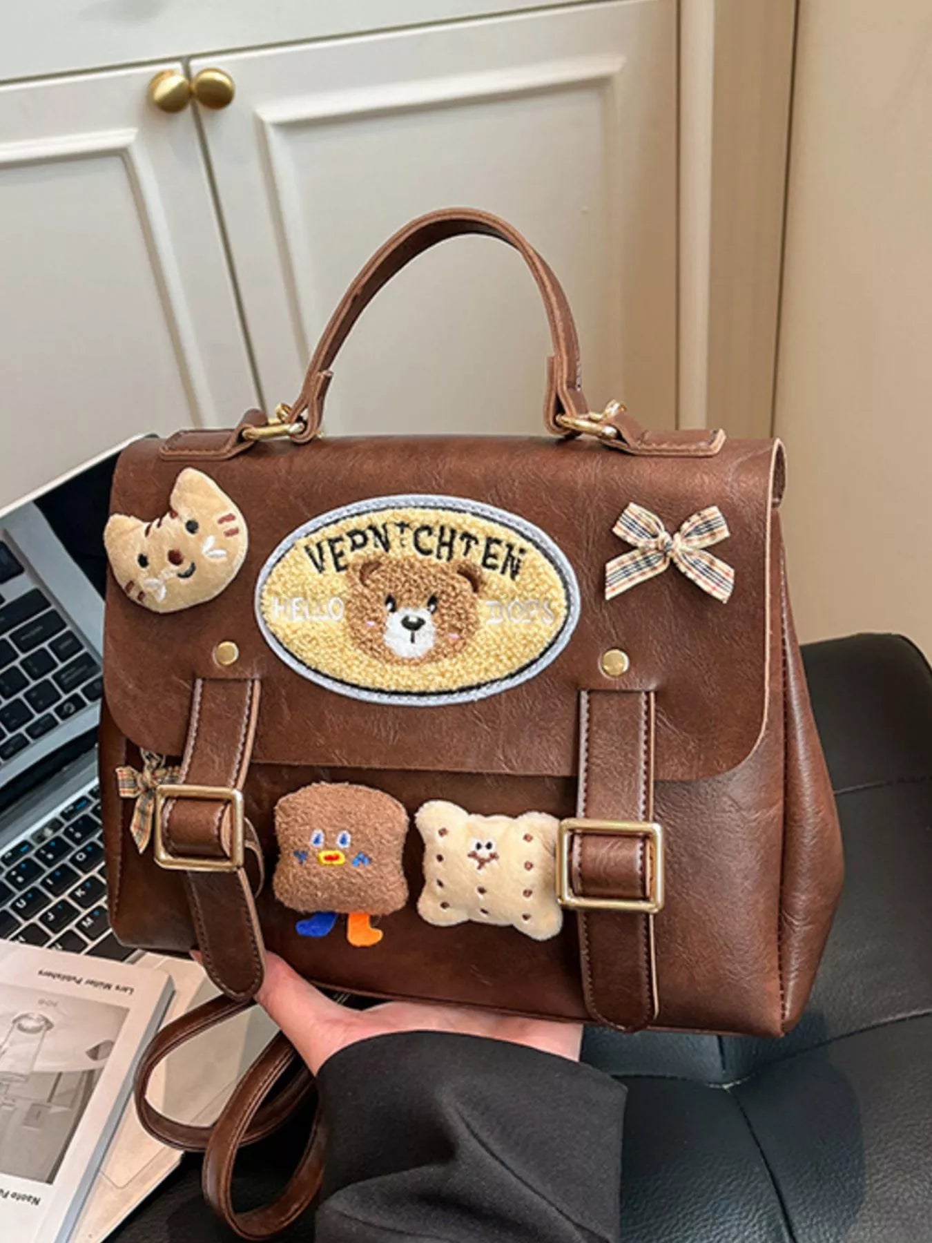 Brown handbag with decorative patches held by a person, with a laptop and newspaper in the background.