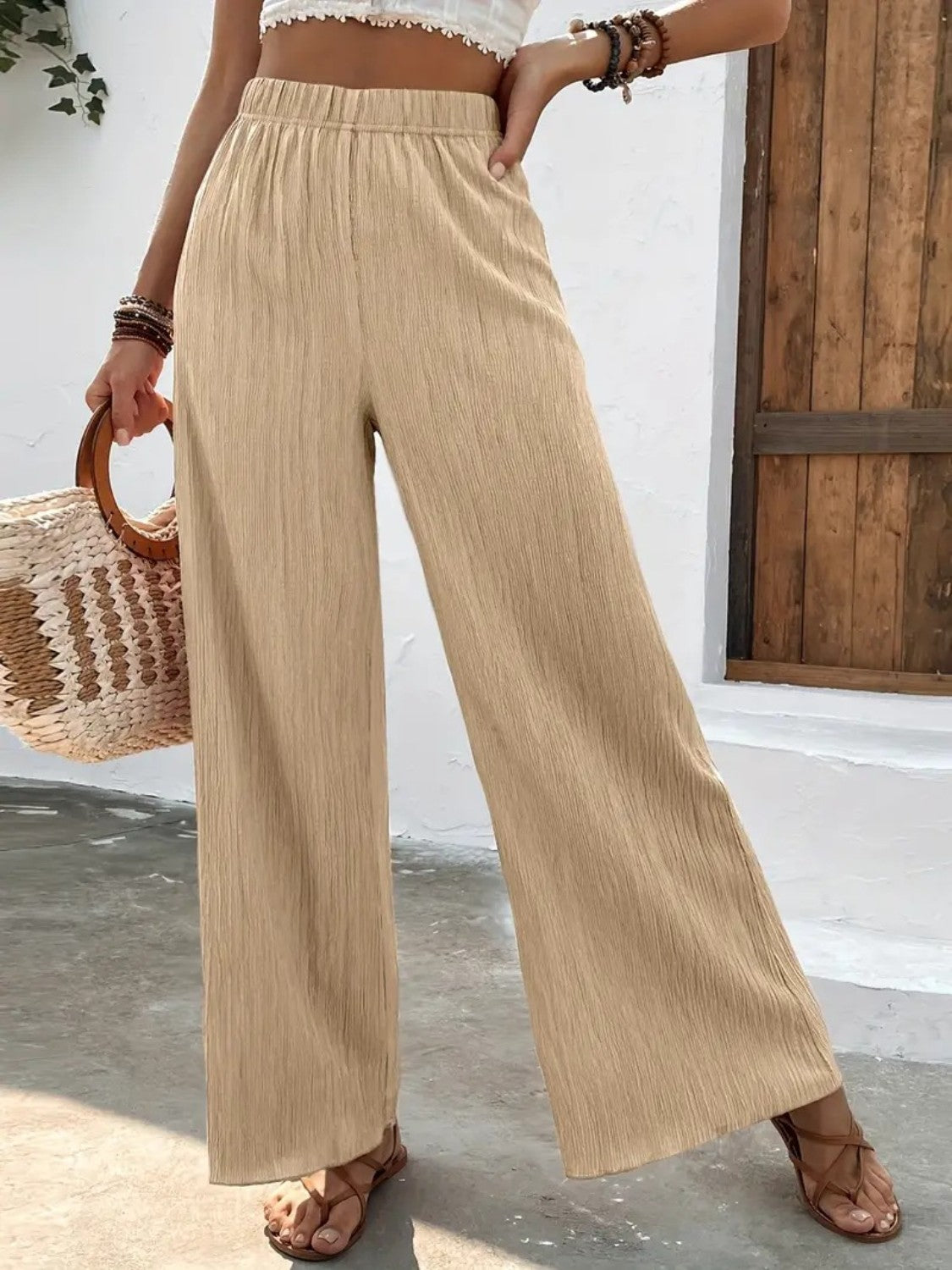 Person wearing beige wide-leg pants holding a woven bag against a white wall.