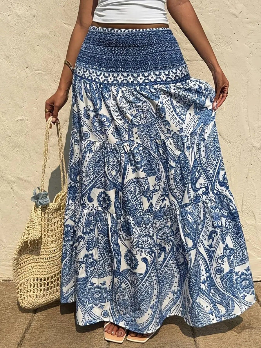 Person wearing a blue and white patterned skirt holding a straw bag against a beige wall.