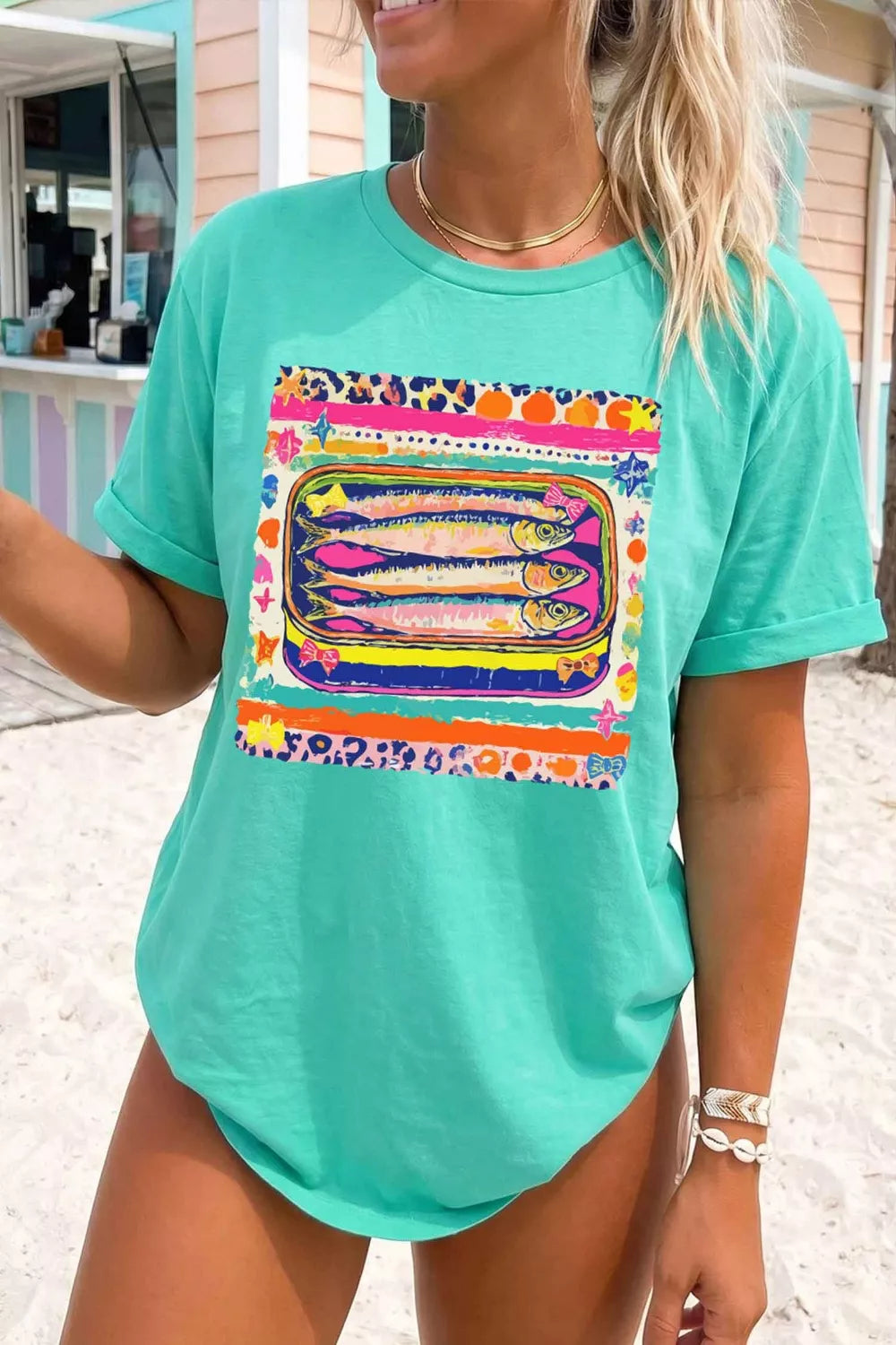 Woman wearing a teal t-shirt with a colorful fish graphic on a beach.