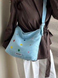 Light blue shoulder bag with colorful polka dots worn by a person in a brown shirt and white pants.