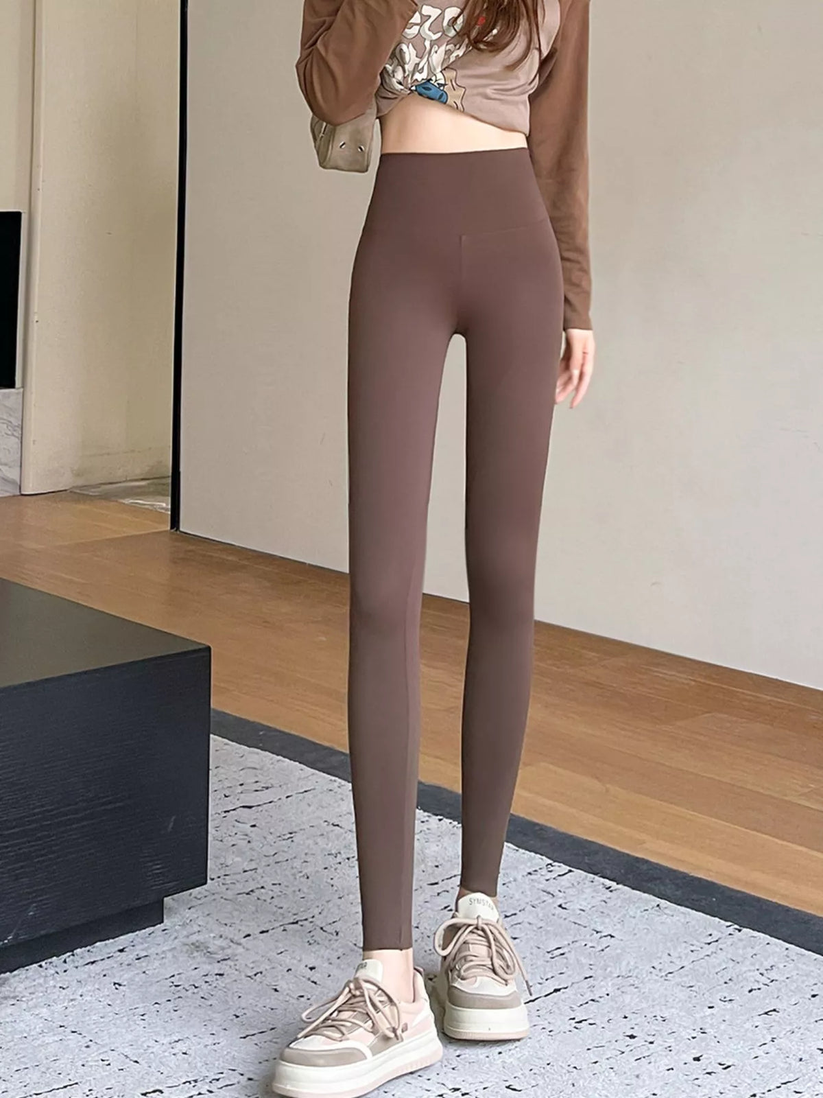 A person wearing high-waisted, form-fitting leggings in a muted taupe-brown color, paired with a cropped long-sleeve top and beige platform sneakers, standing indoors on a patterned rug.