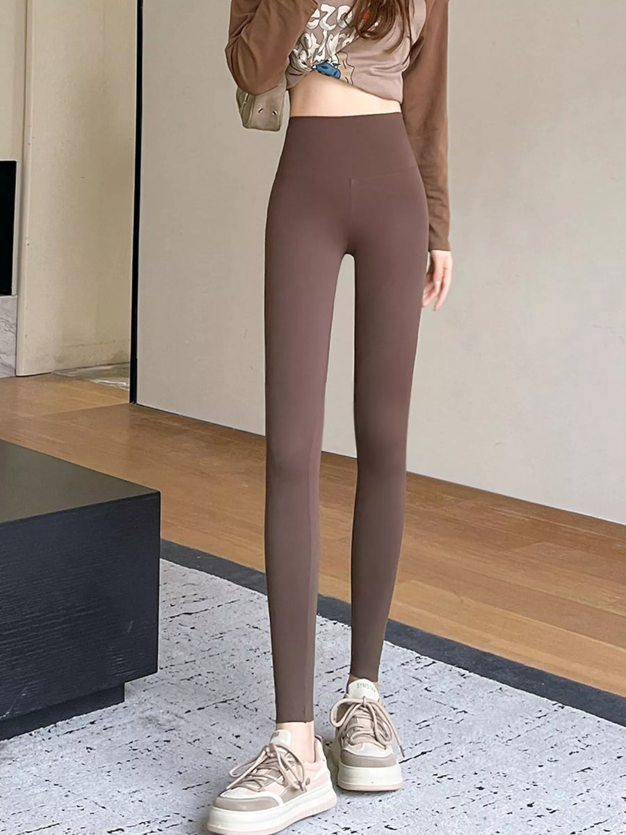 A person wearing high-waisted, form-fitting leggings in a muted taupe-brown color, paired with a cropped long-sleeve top and beige platform sneakers, standing indoors on a patterned rug.