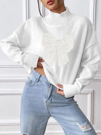 Person wearing a white sweater and light blue jeans against a white wall.