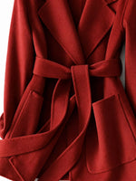 A close-up of a red belted coat or wrap jacket with wide lapels and front patch pockets, shown against a plain light background.