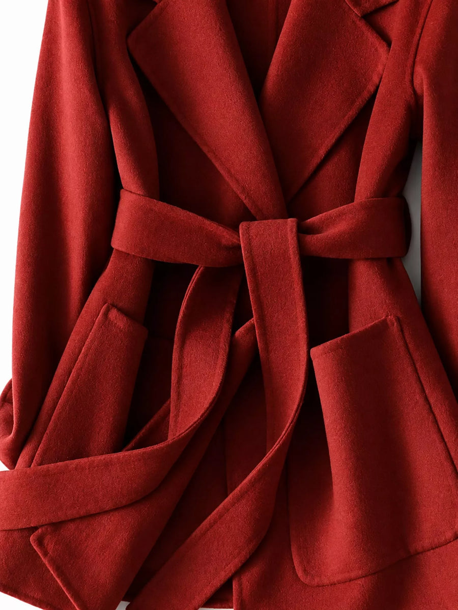 A close-up of a red belted coat or wrap jacket with wide lapels and front patch pockets, shown against a plain light background.