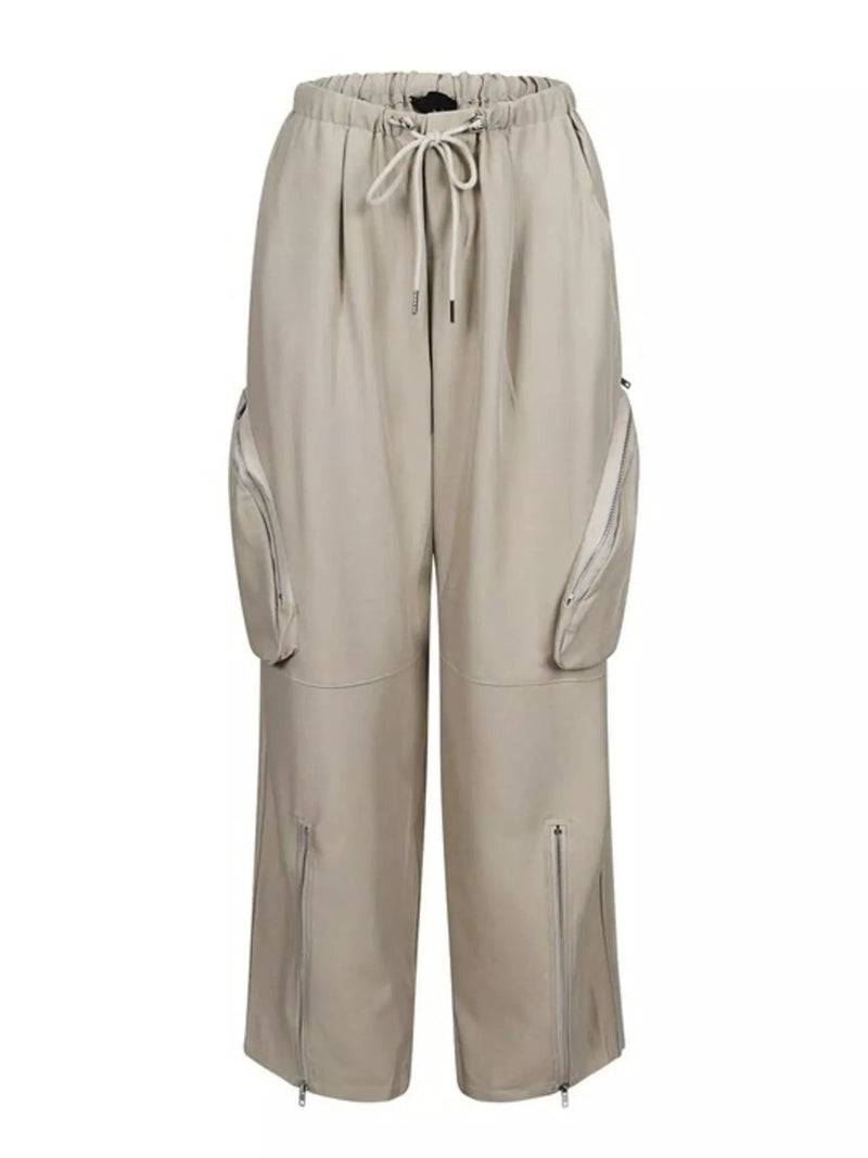 A pair of wide-leg cargo pants in a light beige or sand color, featuring an elastic waistband with a drawstring, large zippered side cargo pockets, and zippered pockets on the lower legs, shown against a plain white background.