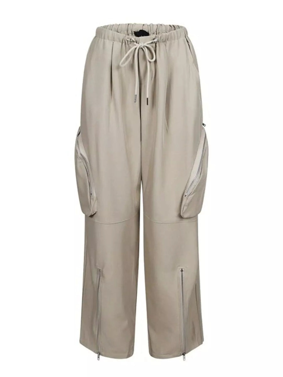 A pair of wide-leg cargo pants in a light beige or sand color, featuring an elastic waistband with a drawstring, large zippered side cargo pockets, and zippered pockets on the lower legs, shown against a plain white background.