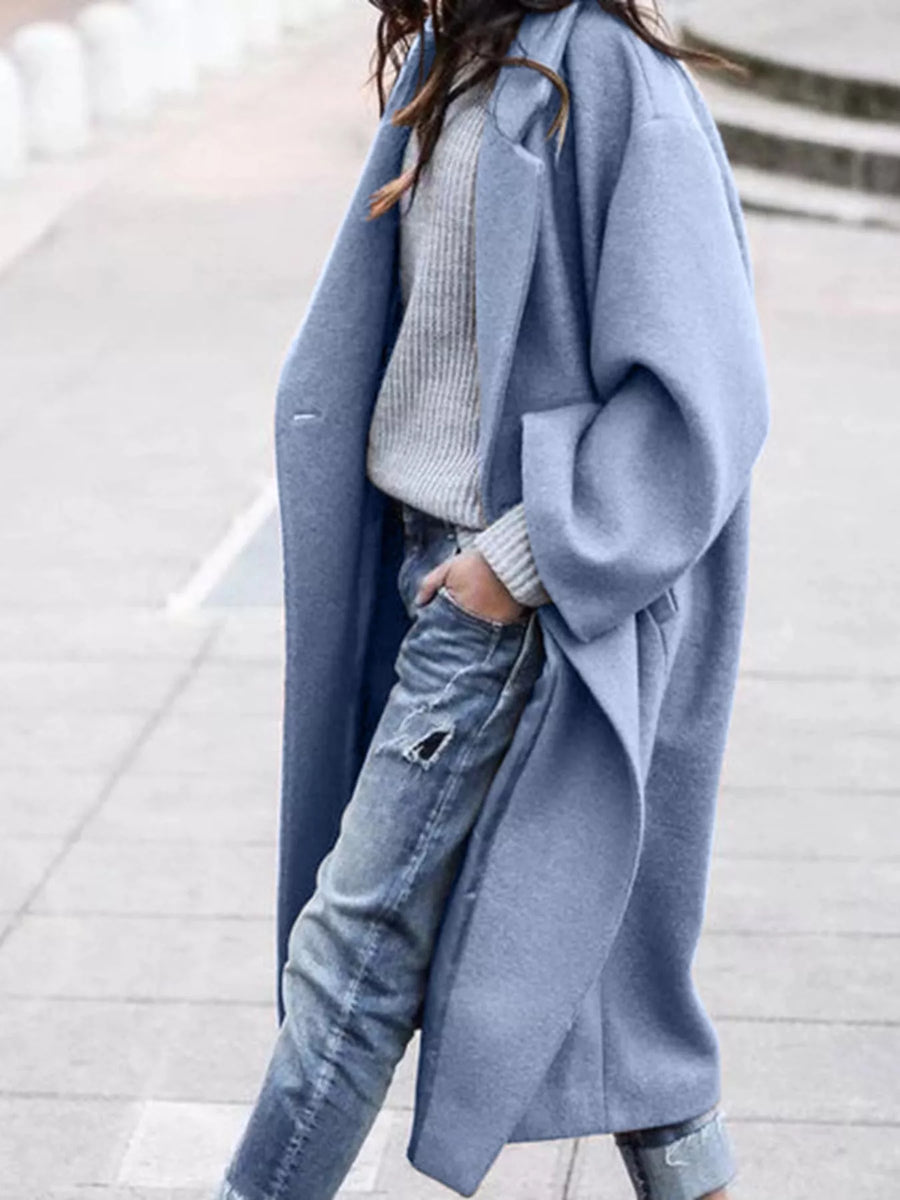 Person wearing a long light blue coat over a gray sweater and jeans on a blurred background