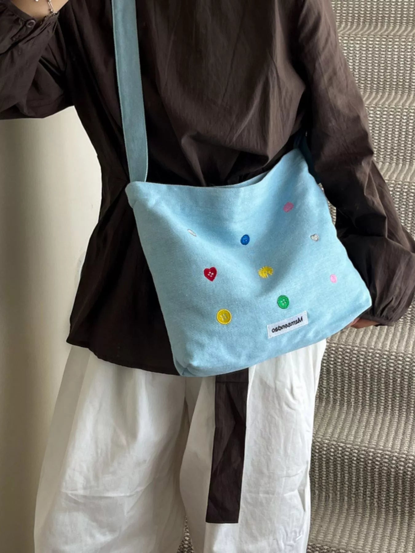 Light blue crossbody bag with colorful embellishments held by a person wearing a brown shirt and white pants.