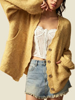A woman wearing a chunky mustard yellow open-front cardigan over a white lace-trim crop top and a light blue denim mini skirt, photographed against a plain light beige studio background.