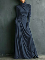 A woman wearing a long-sleeve, high-neck, floor-length navy blue dress with gathered draping at the waist, standing against a dark textured wall.