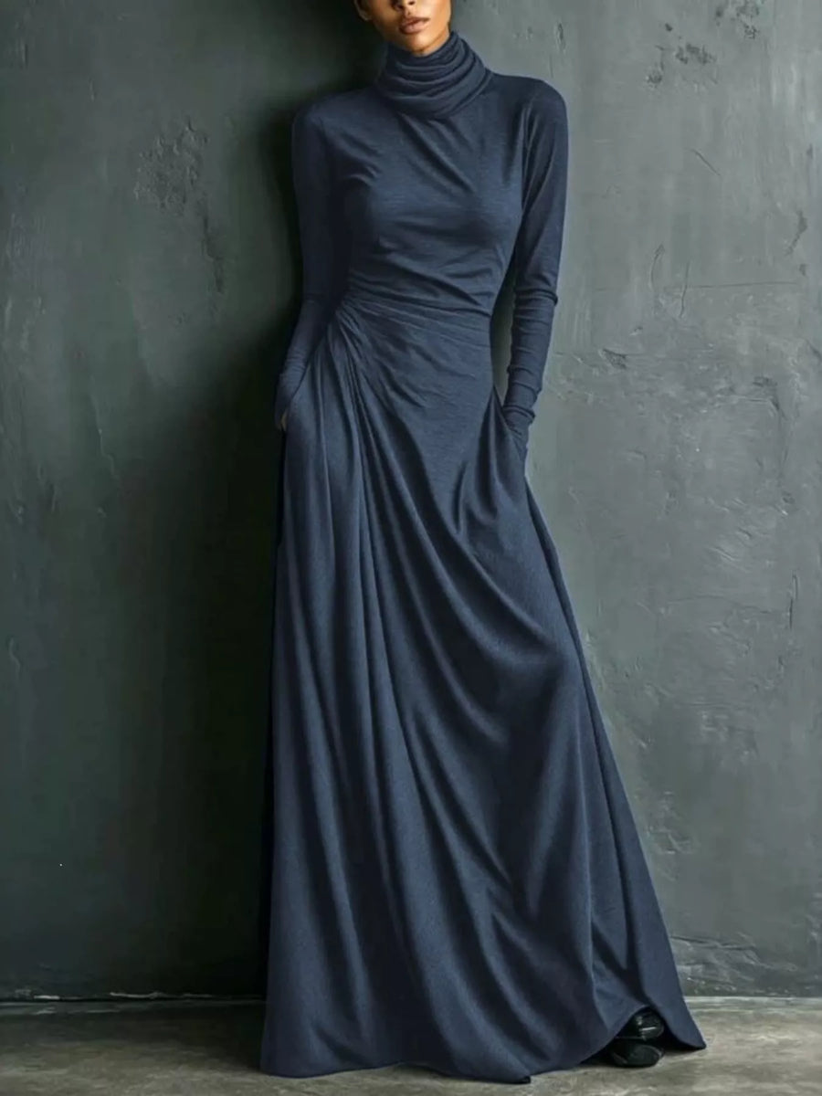 A woman wearing a long-sleeve, high-neck, floor-length navy blue dress with gathered draping at the waist, standing against a dark textured wall.