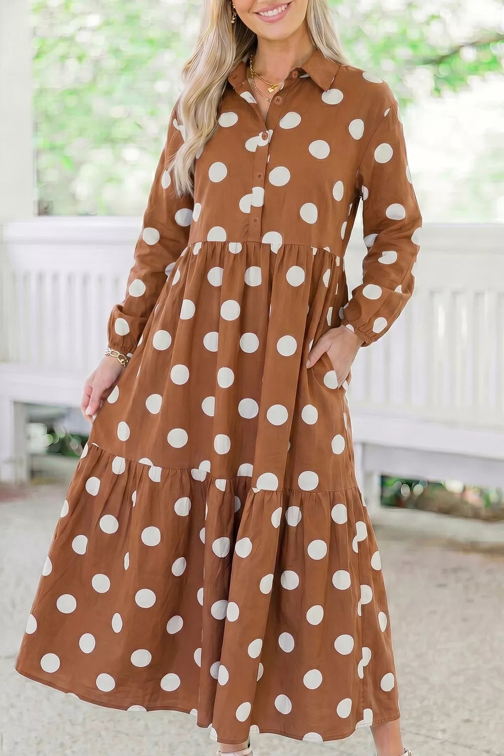 A woman wearing a brown long-sleeve button-up dress with large white polka dots, standing in a bright, airy indoor setting with a white bench in the background.