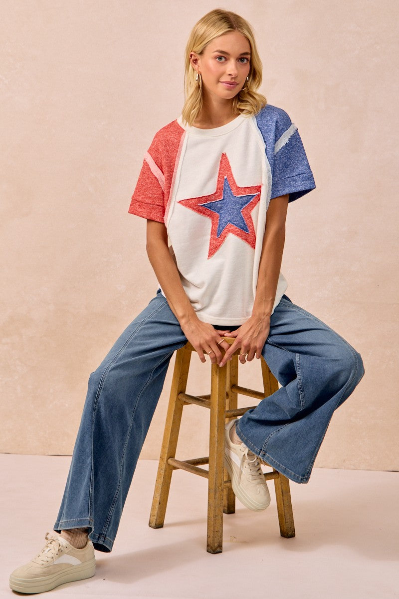 Woman sitting on a stool wearing a colorful star t-shirt and blue jeans against a beige background