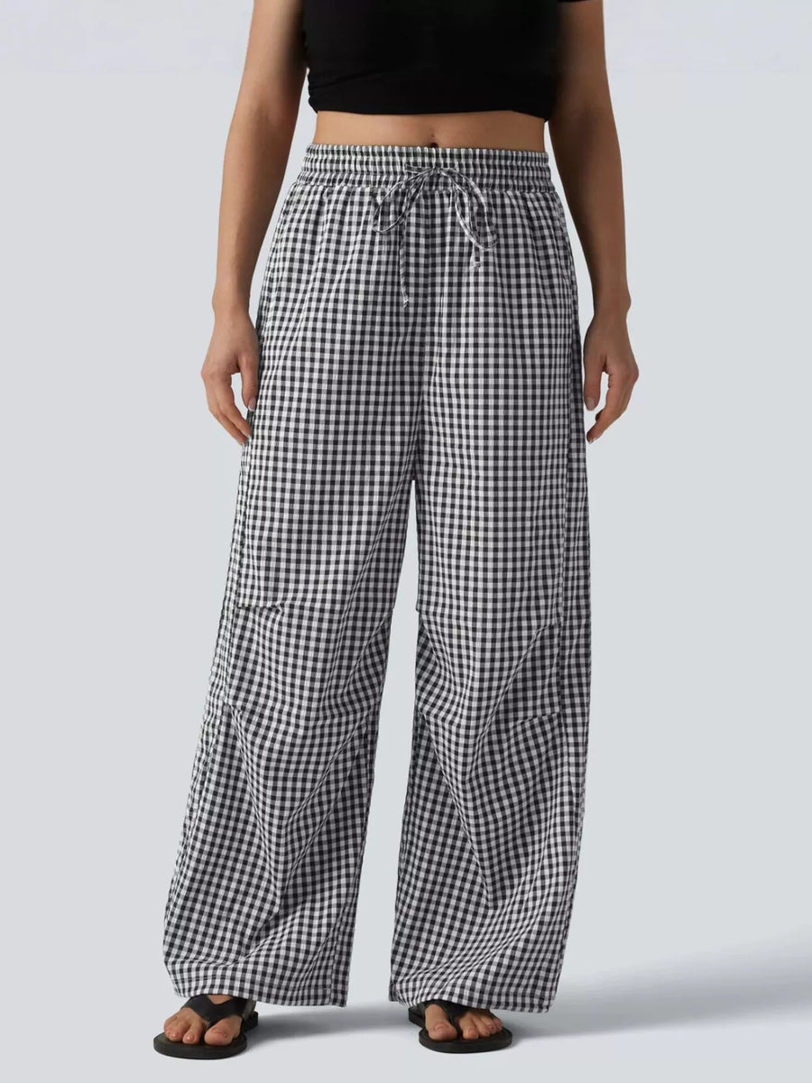 A person wearing loose black-and-white gingham wide-leg pants with an elastic drawstring waist, shown from the chest down against a light gray studio background.