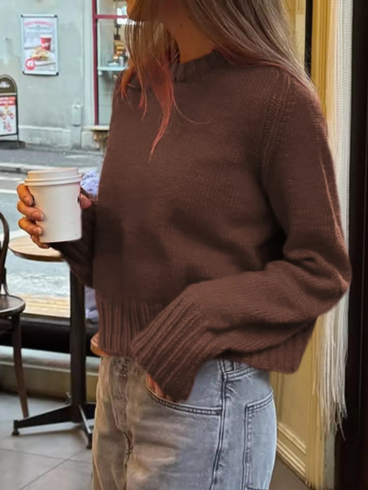 Person wearing a brown sweater holding a coffee cup in an urban setting.