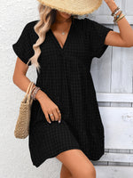 Woman wearing a black checkered dress with a straw hat and holding a woven bag on a wooden floor.