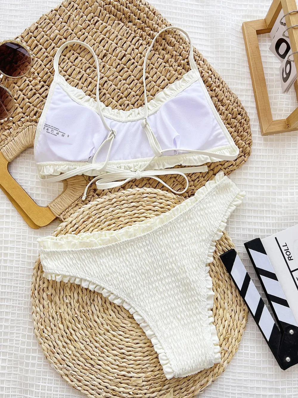 White bikini set on a woven mat with sunglasses and a clapperboard in the background.