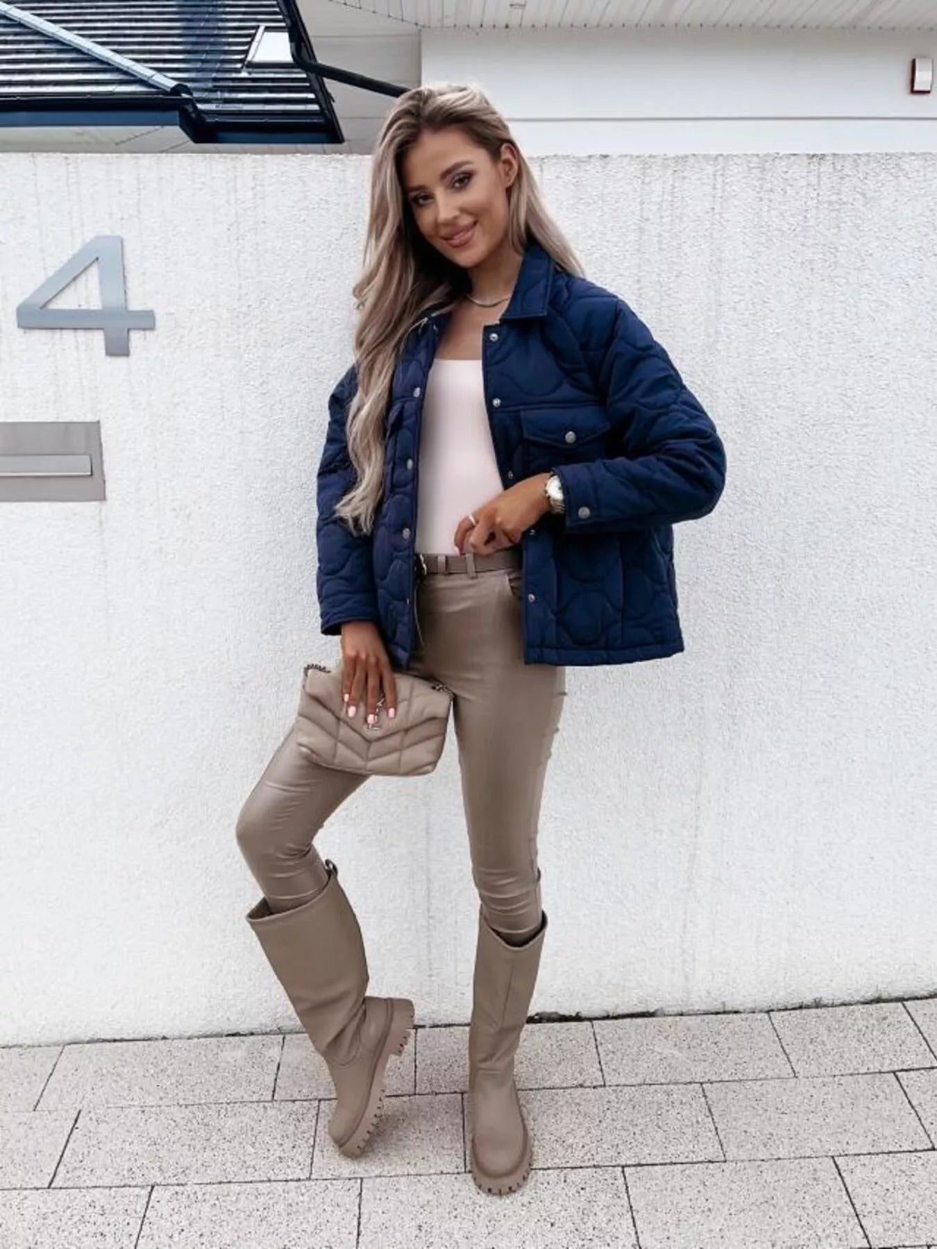 Woman wearing a navy puffer jacket, beige pants, and boots standing against a white wall.