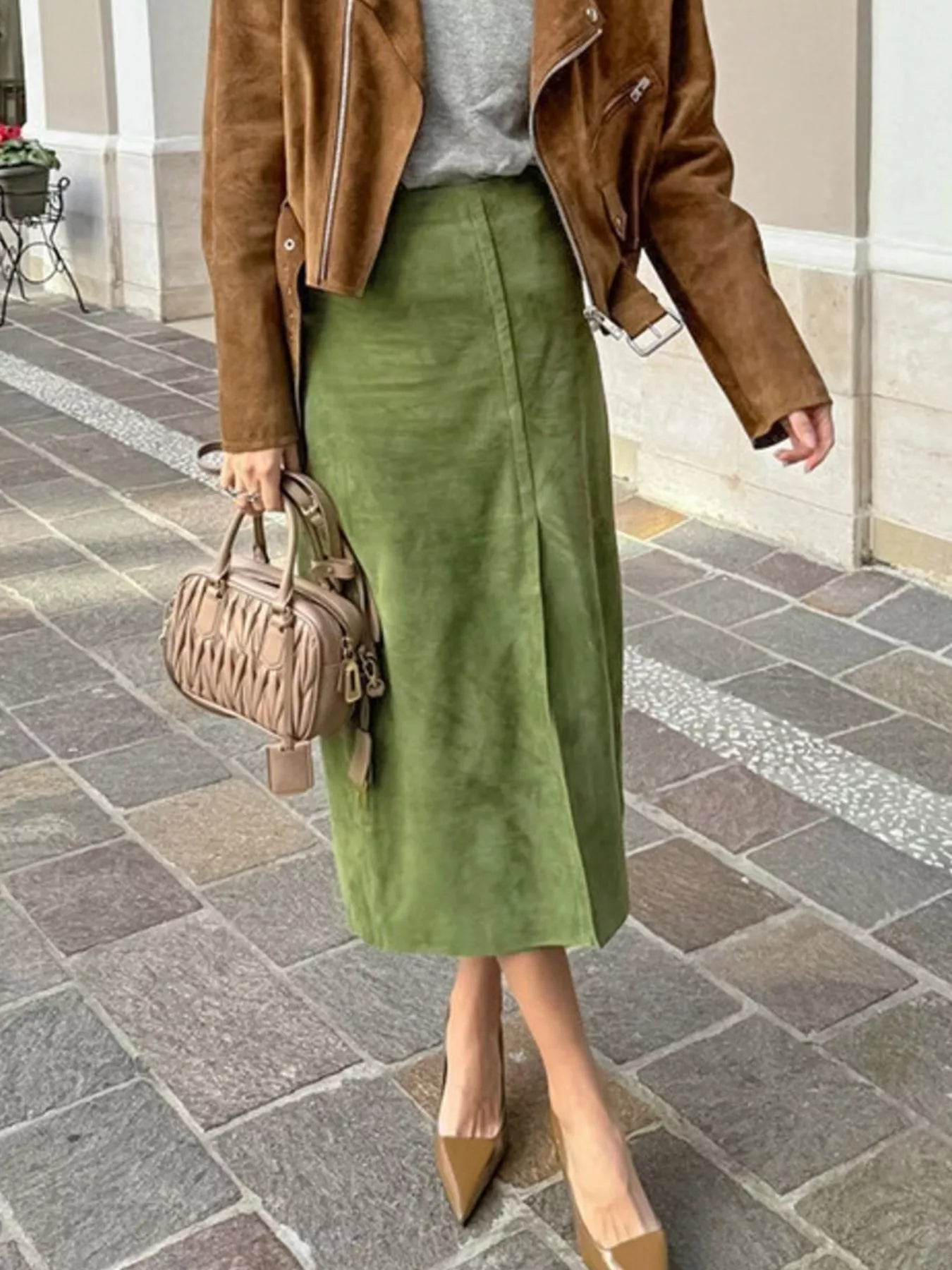 Person wearing a brown jacket, green skirt, and beige shoes on a stone pavement.