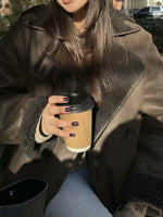 A person wearing a brown shearling-lined coat and holding a takeaway coffee cup outdoors.