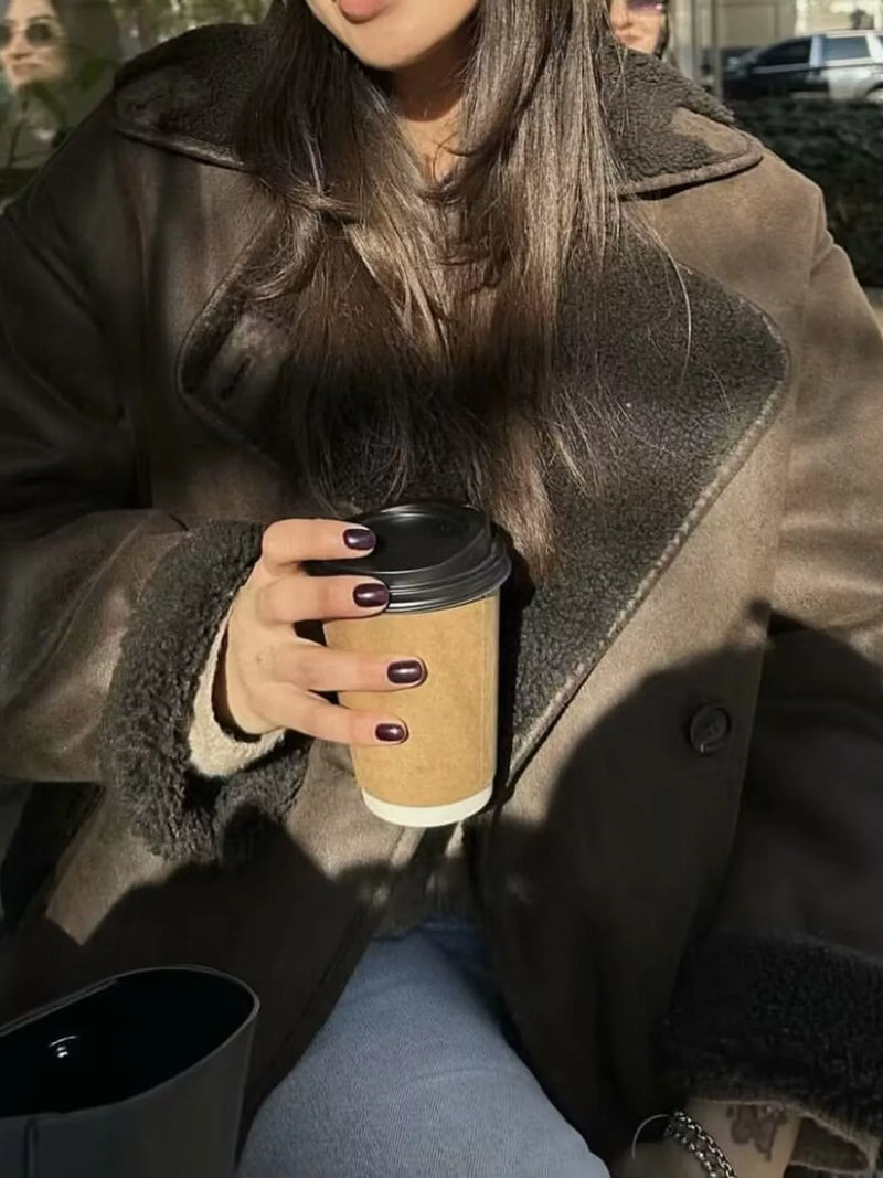 A person wearing a brown shearling-lined coat and holding a takeaway coffee cup outdoors.