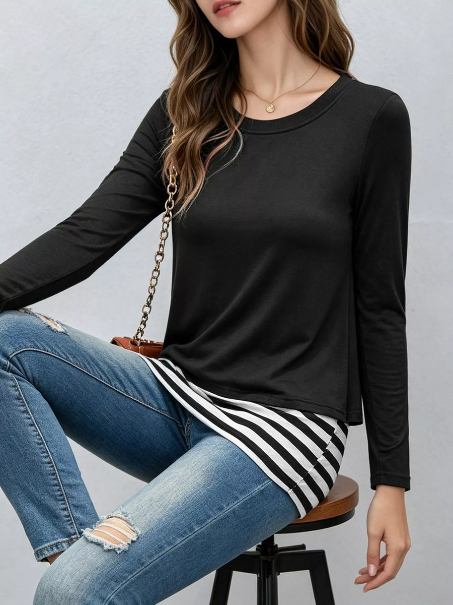 A woman seated on a stool wearing a fitted black long-sleeve top, blue distressed jeans, and a black-and-white striped skirt, with a small brown crossbody bag with a gold chain.