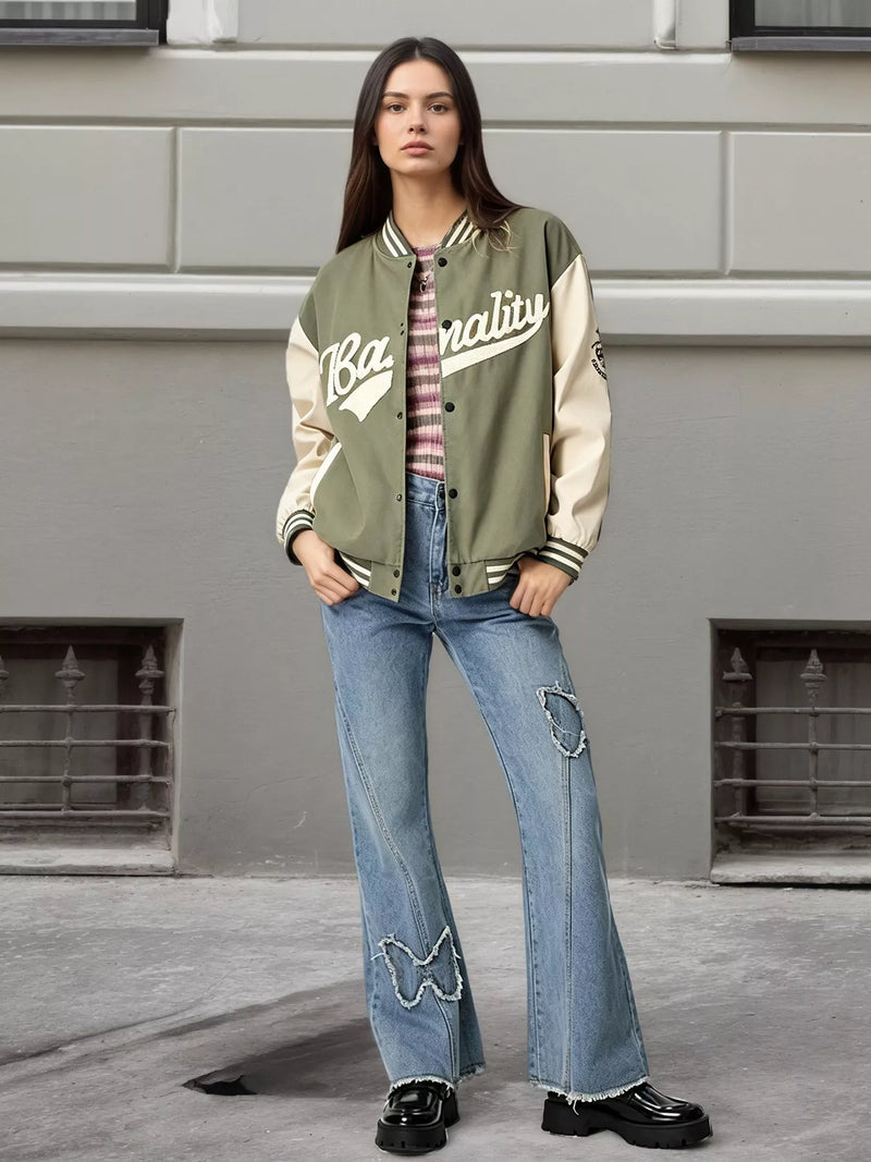 A woman stands outdoors in front of a gray building wall wearing a varsity-style jacket with olive green and cream panels, a striped shirt underneath, light blue distressed flare jeans, and black shoes.