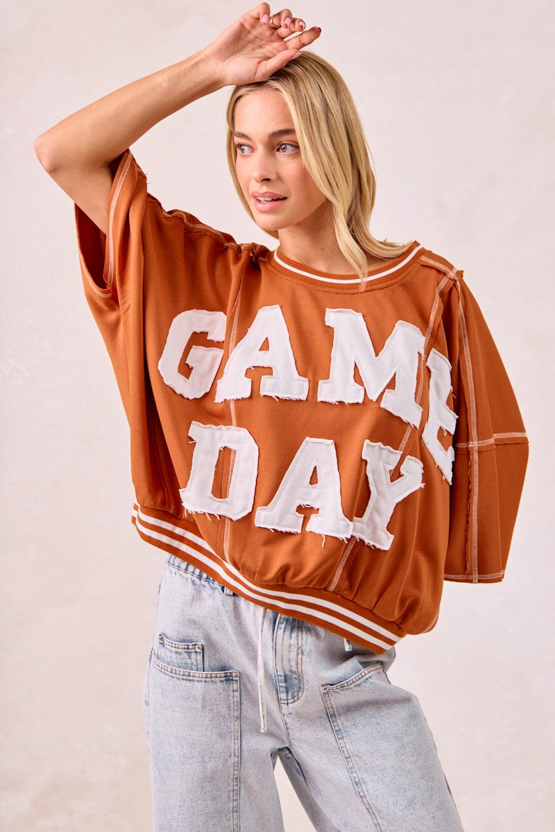 Woman wearing a brown 'GAME DAY' shirt with light blue jeans on a beige background
