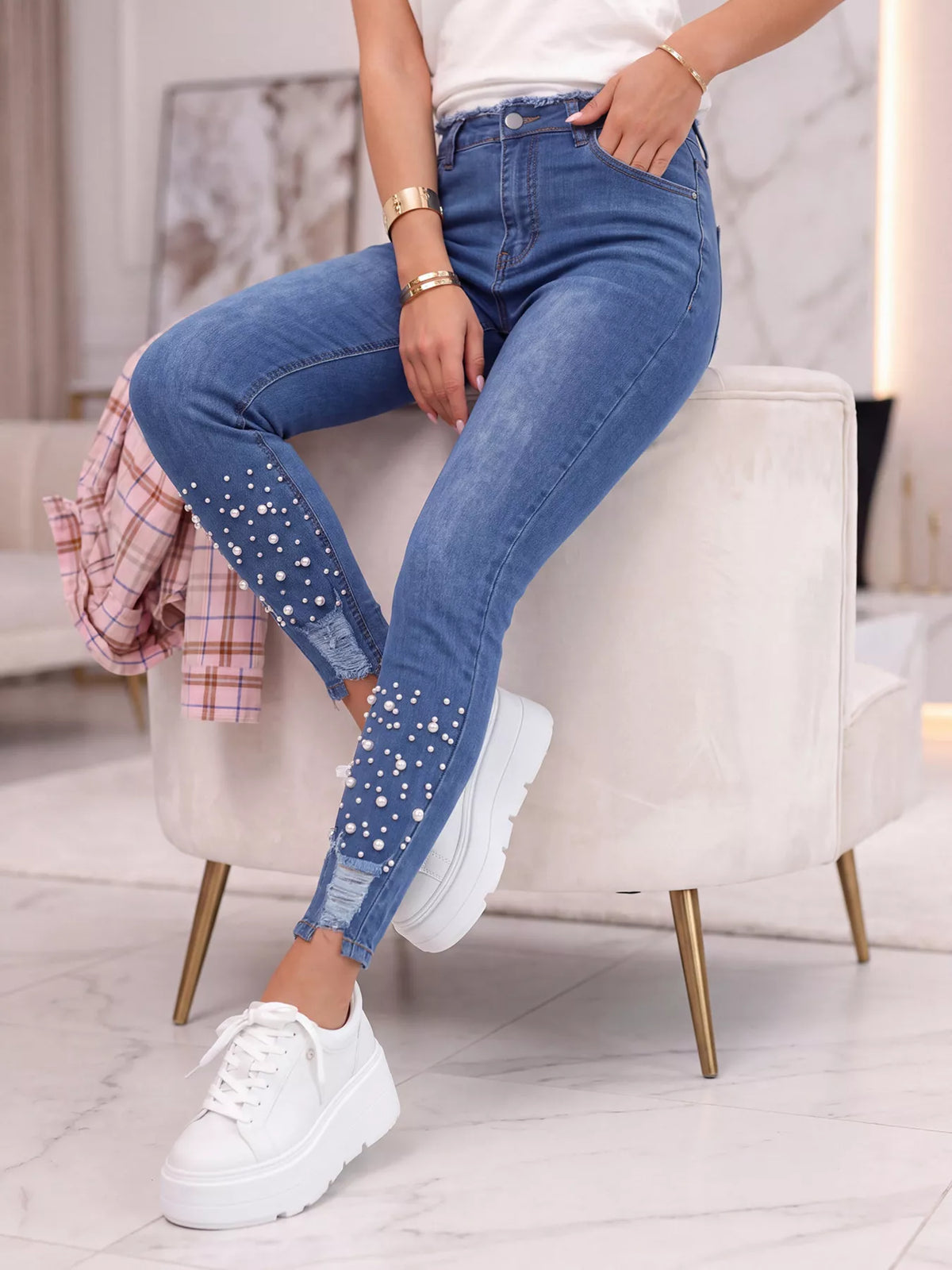 A person seated on a white upholstered chair wearing blue skinny jeans with rhinestone embellishments near the lower legs, a white top, and white platform sneakers in a bright indoor room.